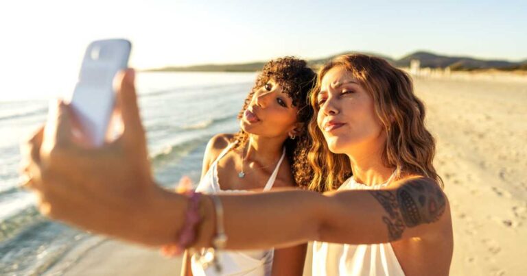 Two cute girls in boho white dress in summer vacation using smartphone taking a selfie making facves standing on sea sand of a tropical beach. Multiracial women travellers having fun with technology
